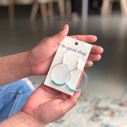 Hand holding a pair of earrings on a 'do good shop' card with a blurred indoor background.