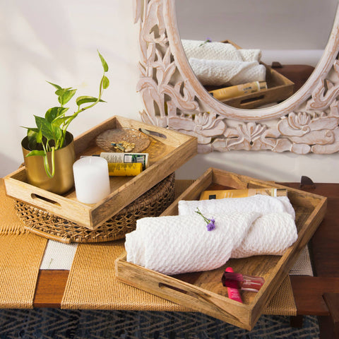 Two wooden serving trays with one featuring a two-tone design, placed on a table with decorative items, a plant, and a mirror in the background.