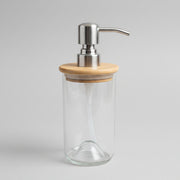 A handmade soap dispenser made of clear glass with a bamboo lid and a pump dispenser on top, placed on a wooden surface.