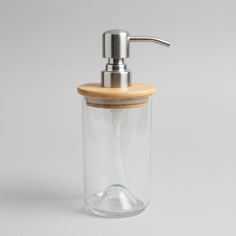 A handmade soap dispenser made of clear glass with a bamboo lid and a pump dispenser on top, placed on a wooden surface.
