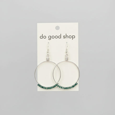 Hand holding a card with earrings labeled 'do good shop' against a wooden background