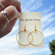 Silver hoop earrings on a 'do good shop' card held against a blue sky with trees.