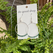 Earrings on a 'do good shop' card hanging from a tree branch with green leaves.