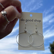 Silver hoop earrings on a 'do good shop' card held against a blue sky with trees.