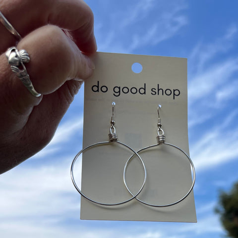 Silver hoop earrings on a 'do good shop' card held against a blue sky with trees.